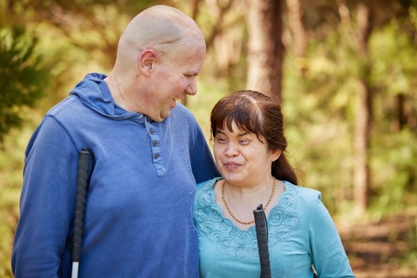 A happy couple standing close together and smiling at each other. It is sunny and there are trees in the background.