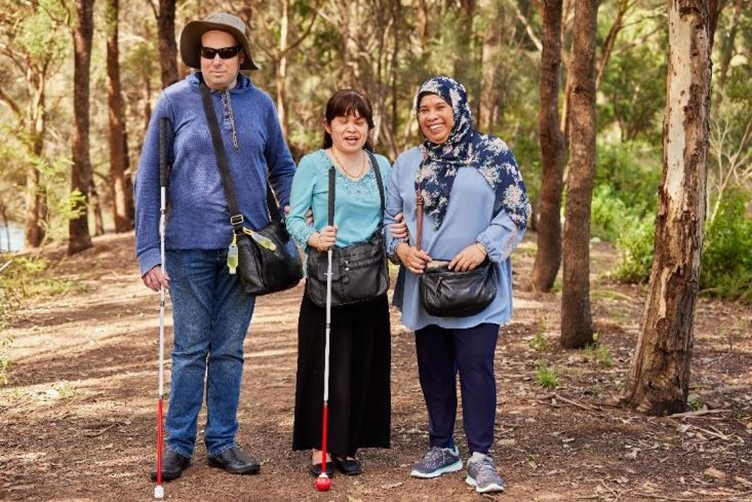 A man and two women standing in a park. They are smiling and surrounded by trees. 