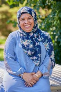 A woman wearing a blue top and floral head scarf. She is smiling while sitting on a bench at the park.