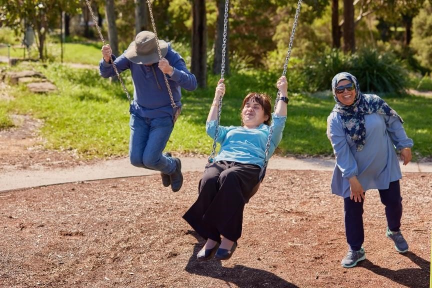 A man and a woman swinging through the air happily on neighbouring swings. A woman stands in the background with a smile on her face. 