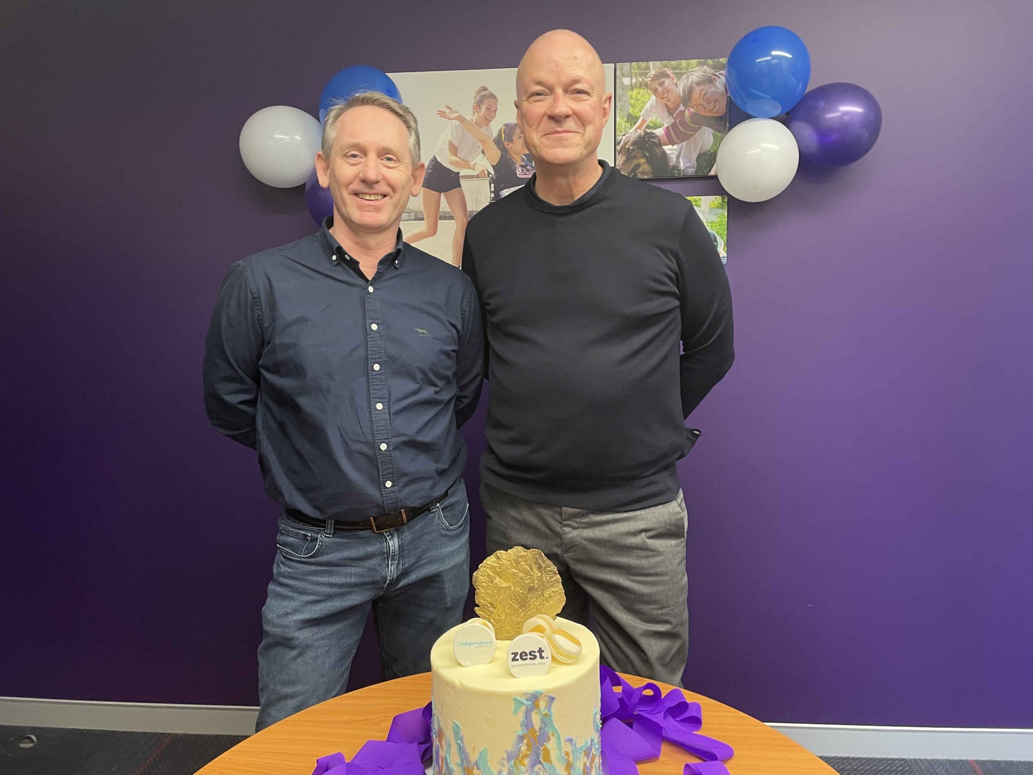 Two men smiling at the camera in front of balloons and purple wall.