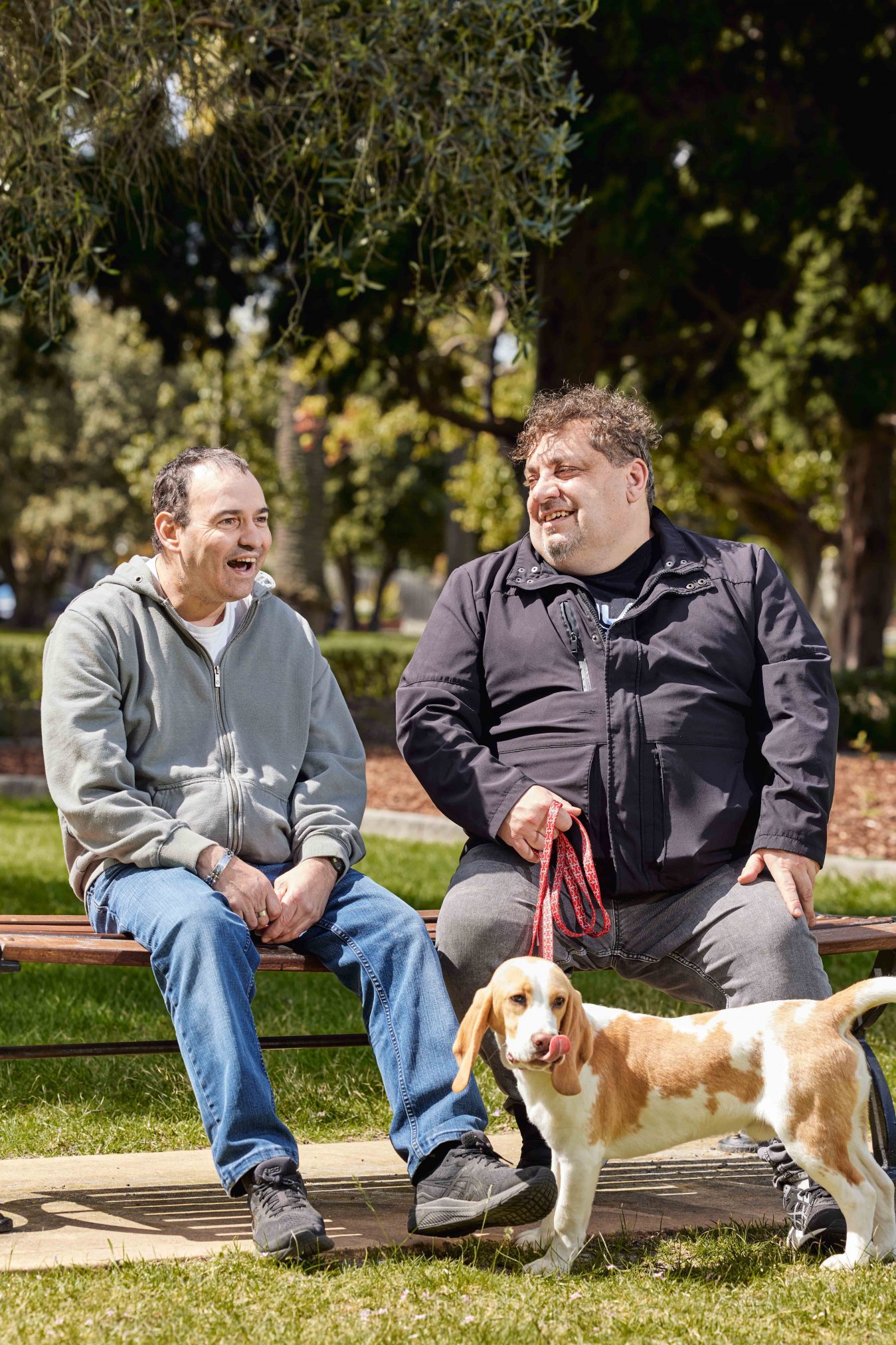 Client and Support Worker laughing together on park bench. The dog is being held on its leash by Support Worker.