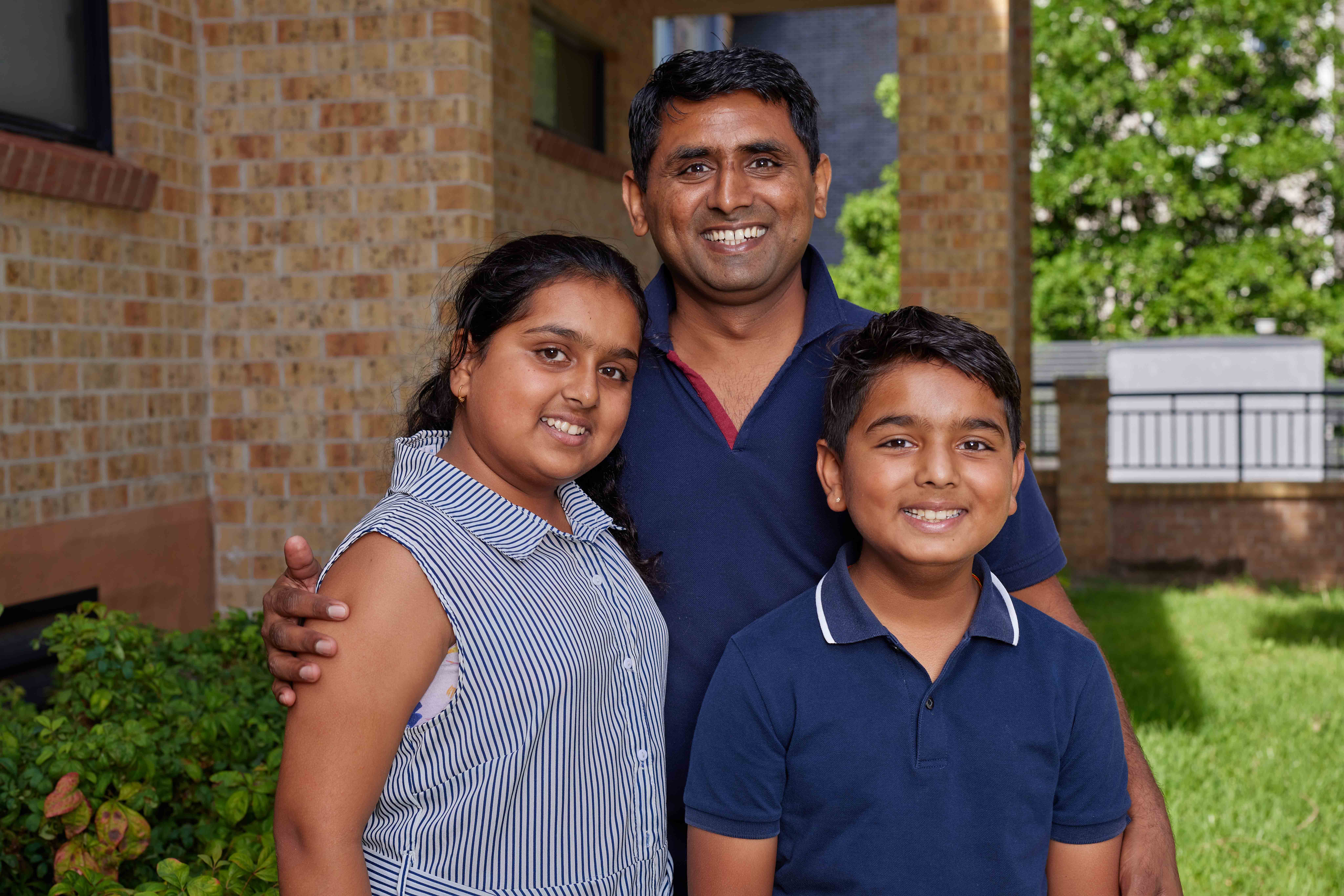 A father standing with his two smiling kids. They are outside and there is a leafy tree in the background.