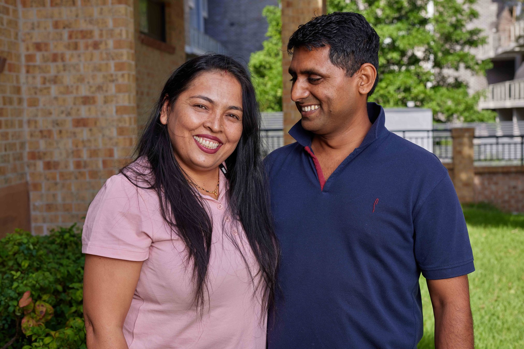 Husband smiling and looking fondly at his wife who is smiling at the camera. They are outside and there is a leafy tree in the background.