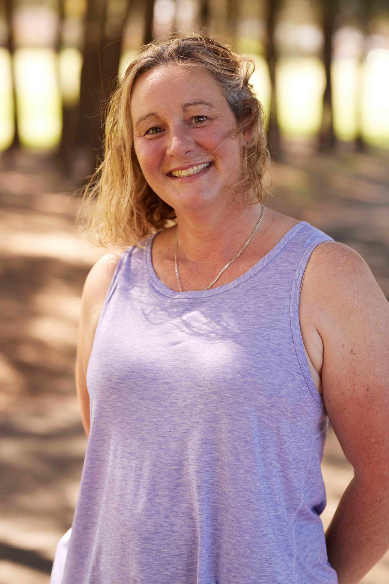 A portrait of a woman, she is smiling in the sun, there are trees in the background.