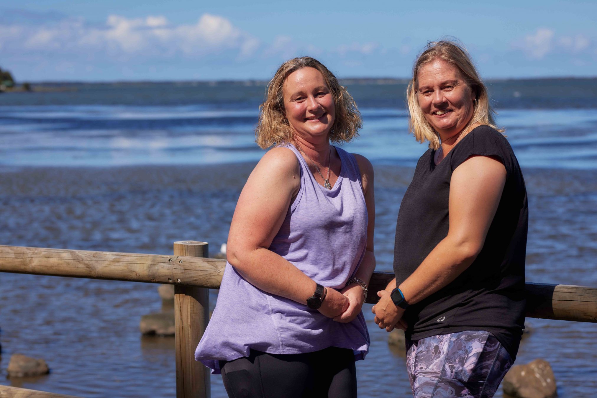 Two women smiling at the camera, there is a lake in the background.