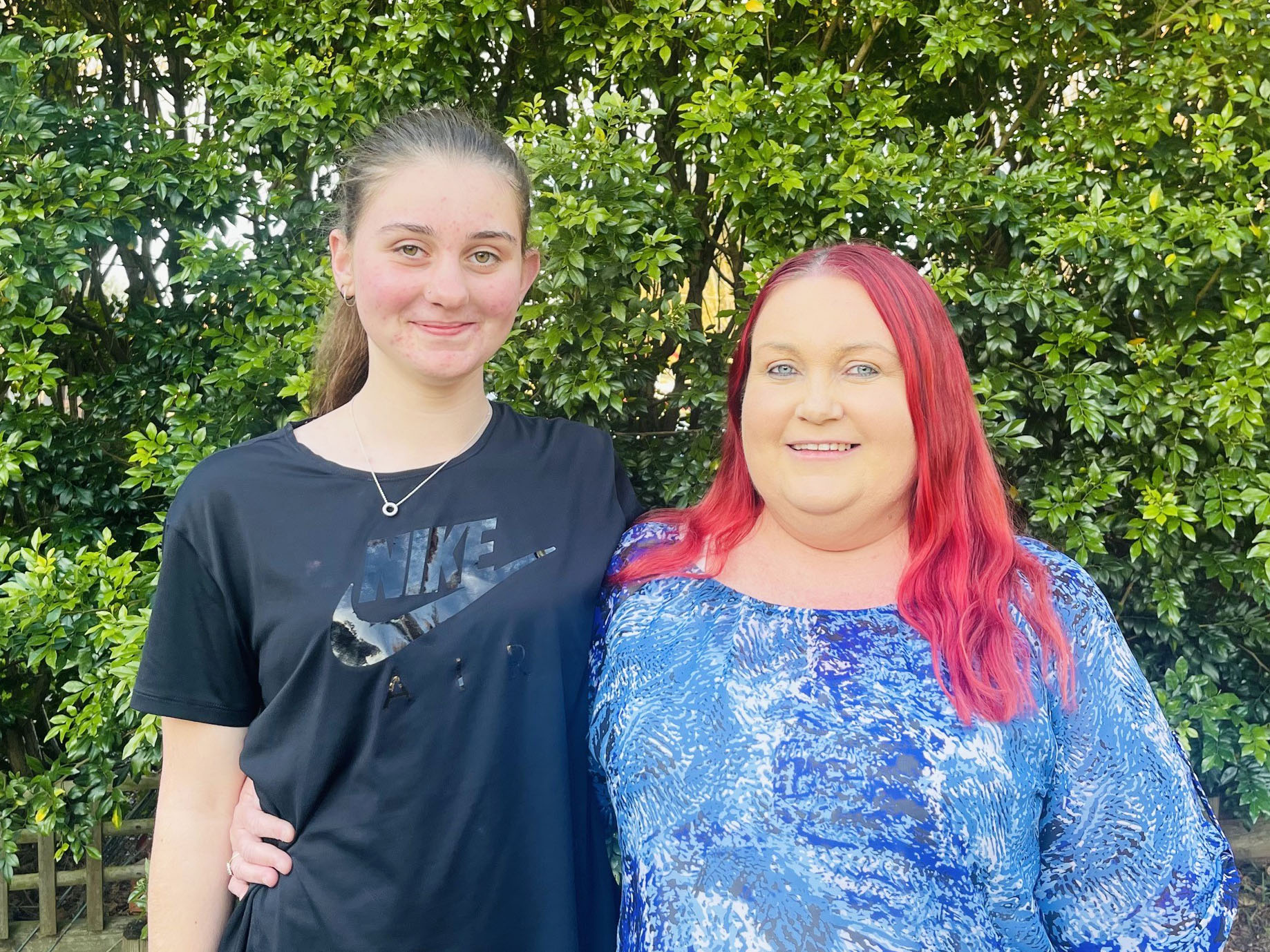 Mother and daughter smiling at camera, green shrub in background.