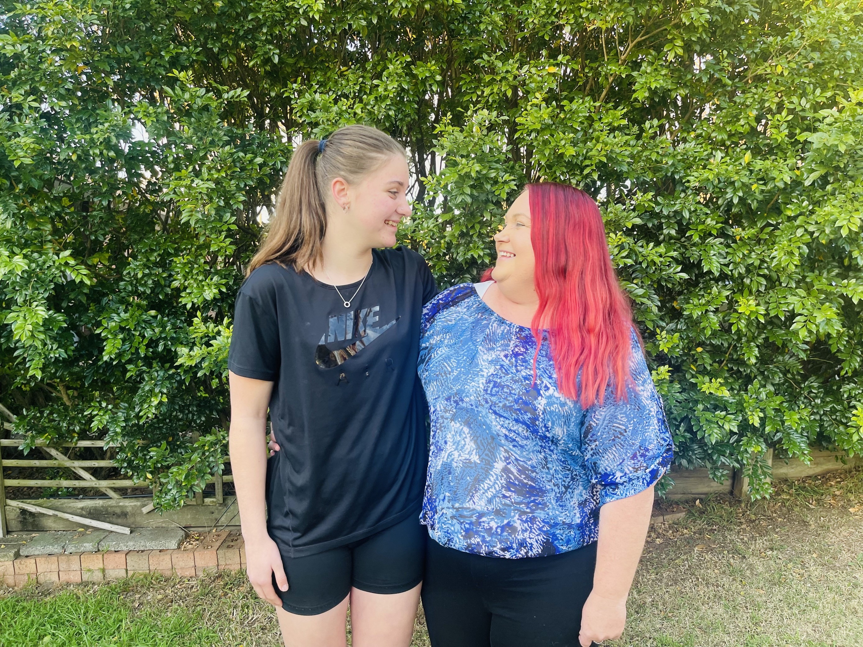 Mother and daughter looking at each other and smiling. They are standing outside in front of shrub.