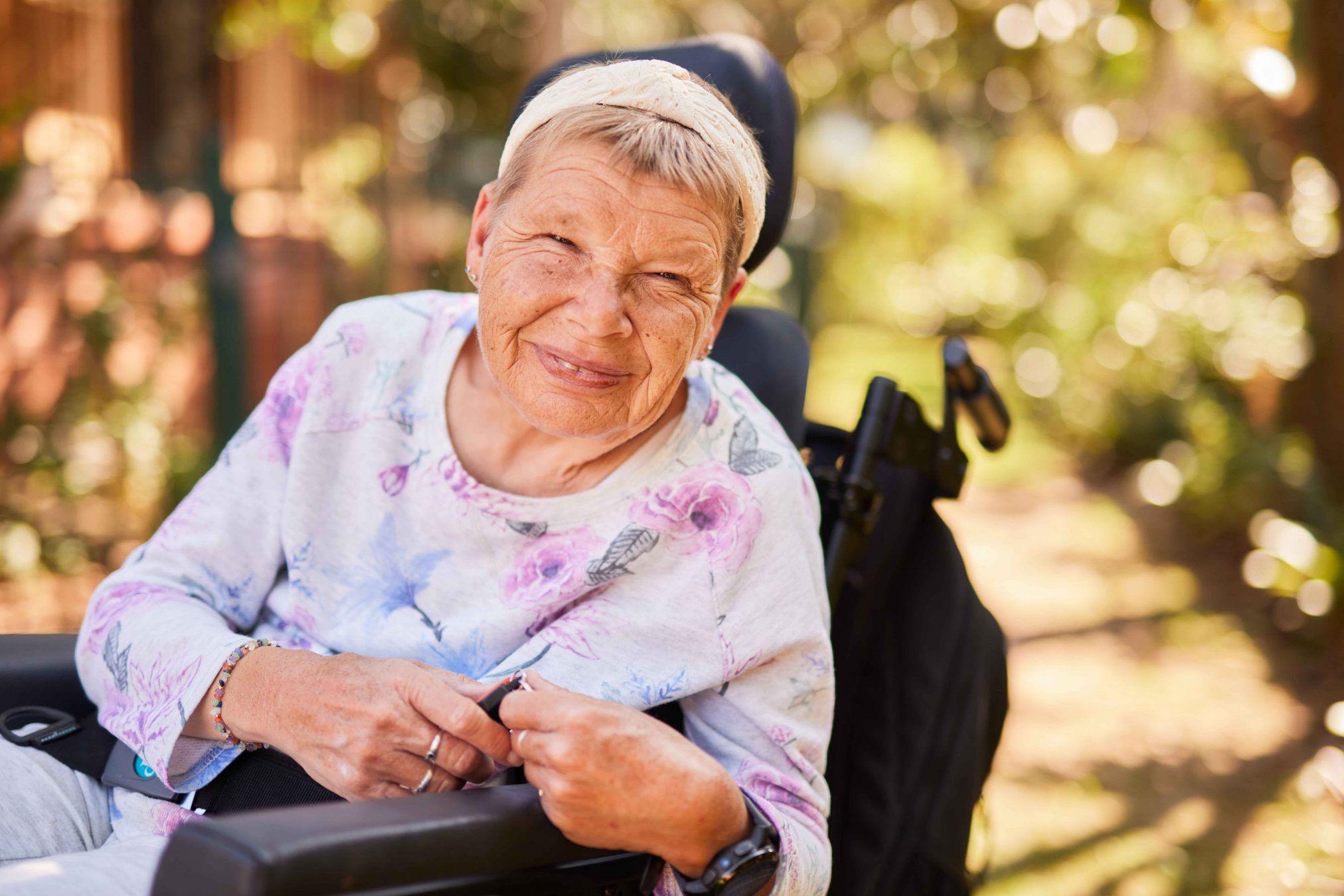 A close up of a woman smiling. She is outside and there is bright dappled light surrounding.