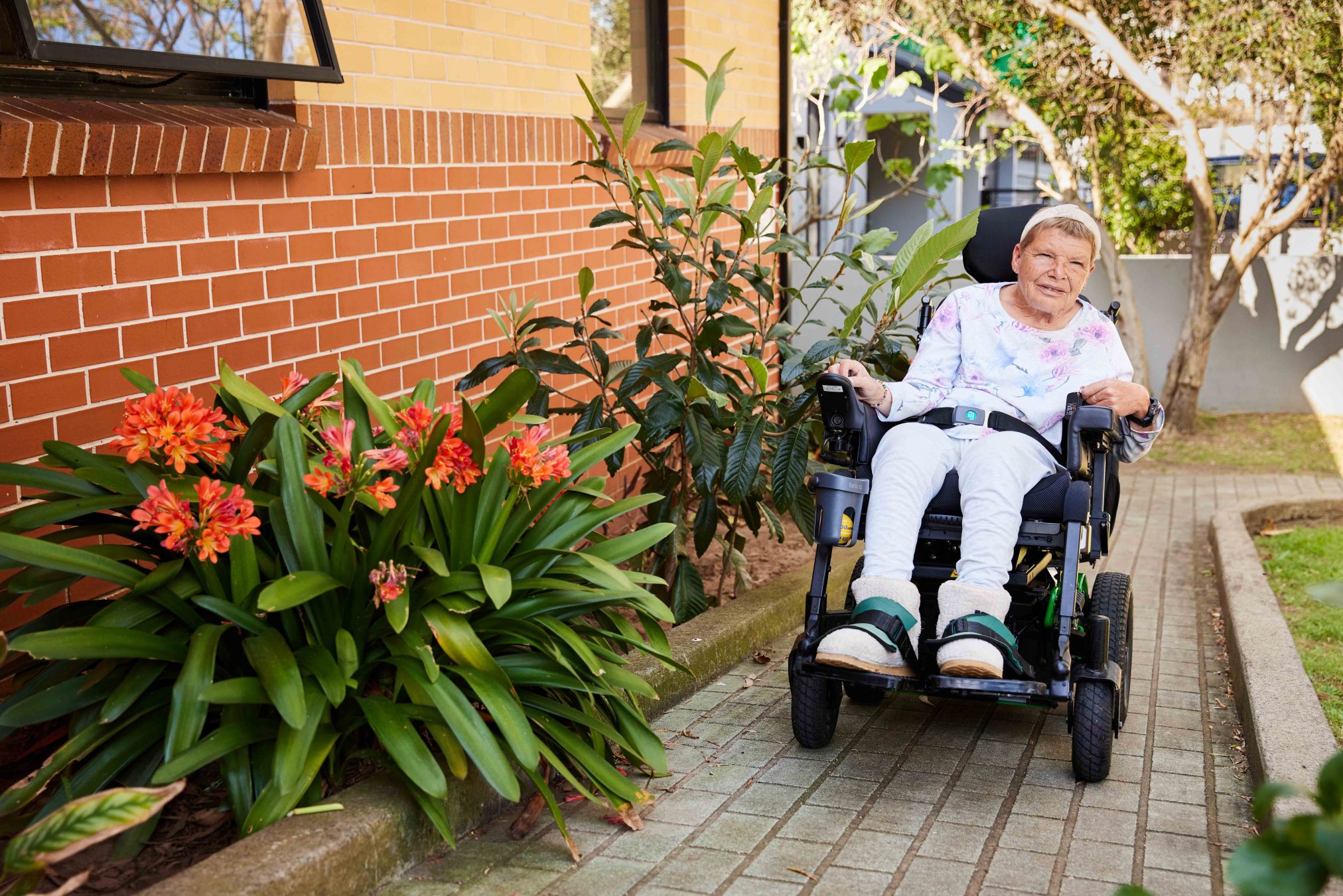 Woman in a powerchair coming up a ramp, smiling. Bright orange flowers surround.