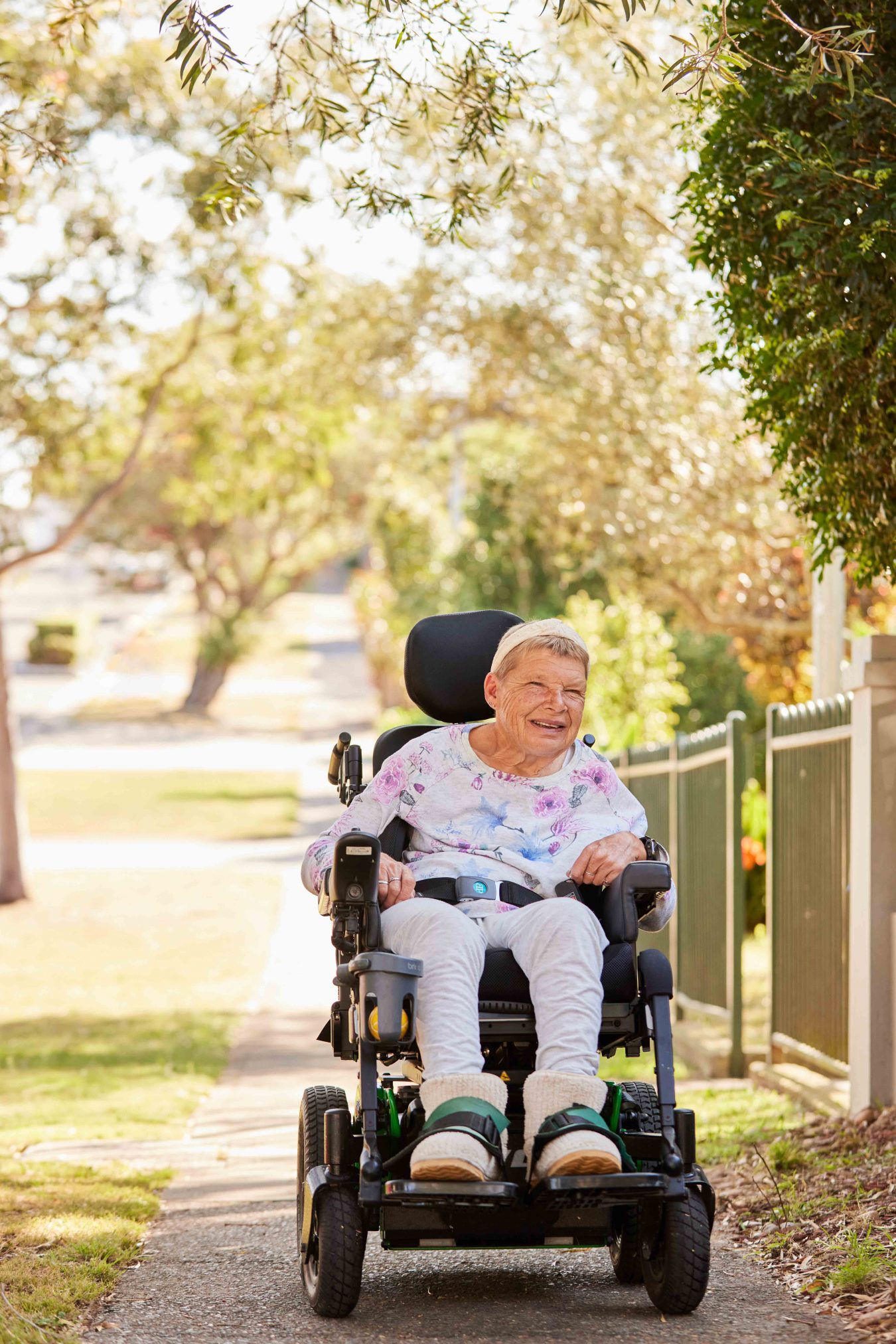 A woman driving along in her powerchair on the footpath. She is smiling and looking off into the distance.