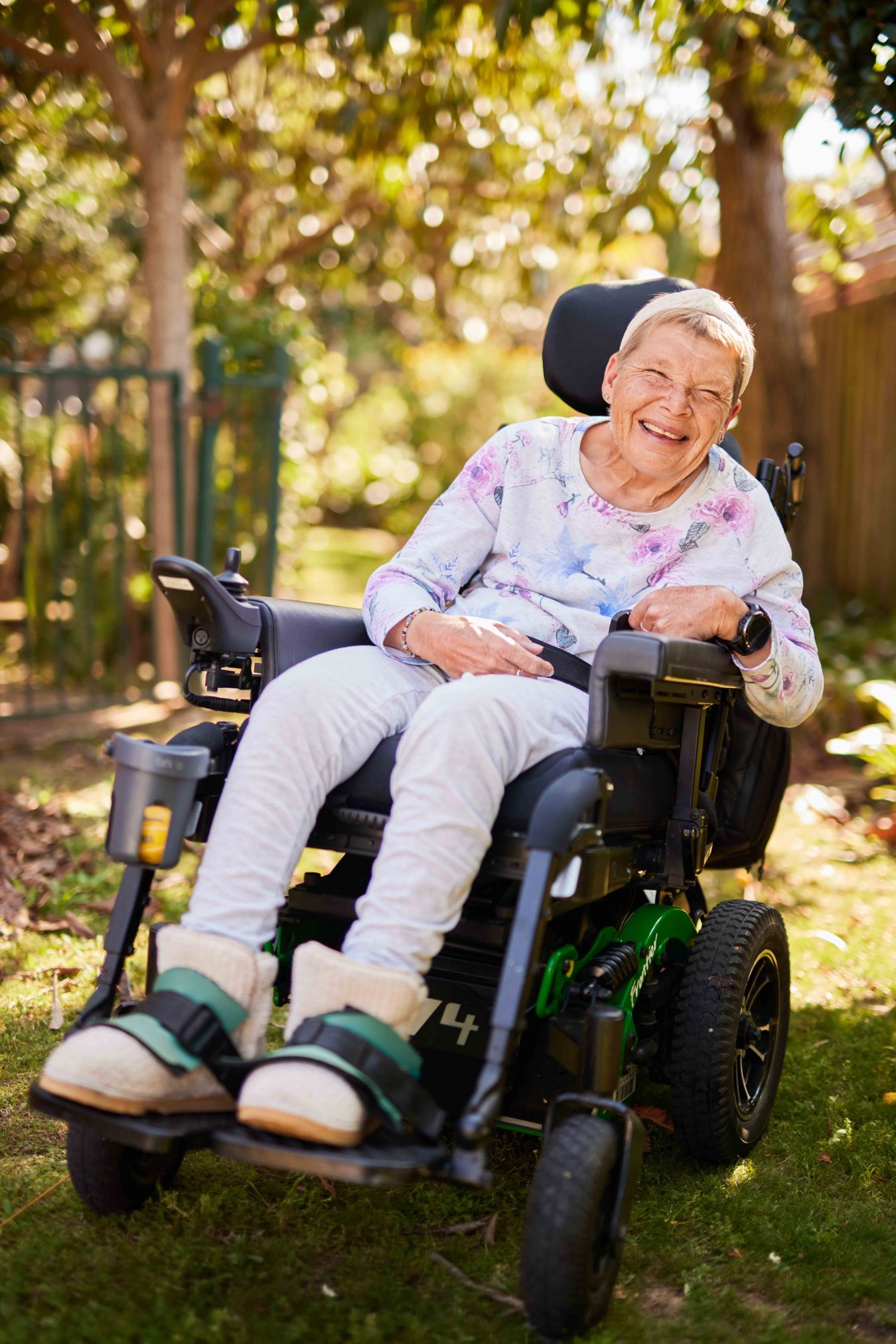 A woman sitting in her powerchair and laughing.. She is sitting outside in a courtyard.